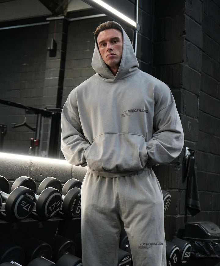 Model wearing a grey HERCSTUDIOS vintage boxy hoodie and matching sweatpants beside a gym dumbbell rack