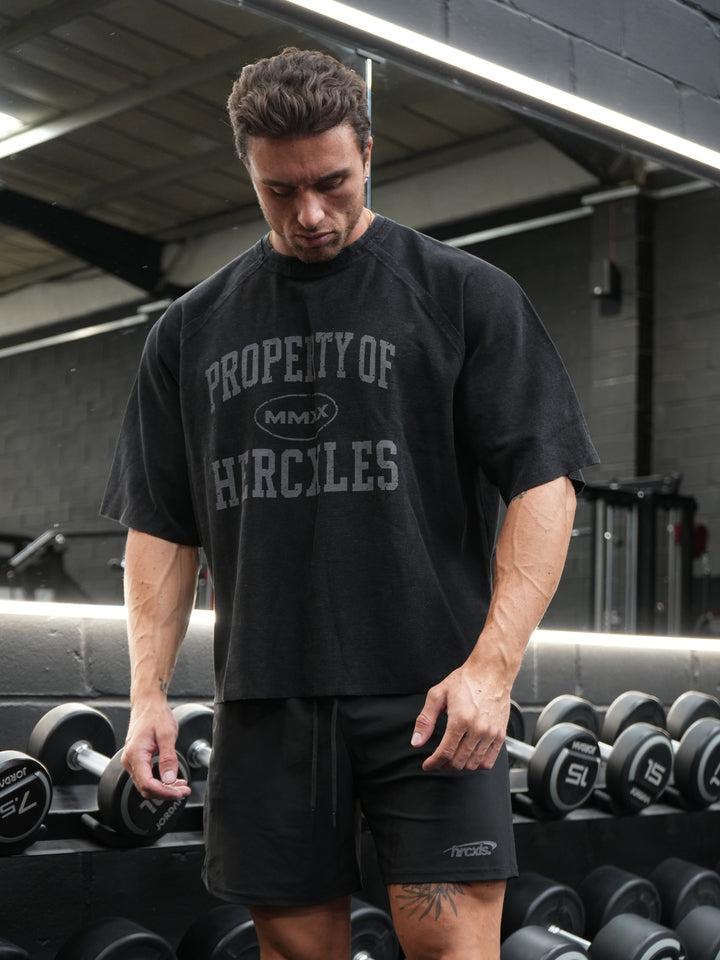 Athlete wearing an oversized black Property of HERCXLES waffle T-shirt and black shorts in front of a dumbbell rack