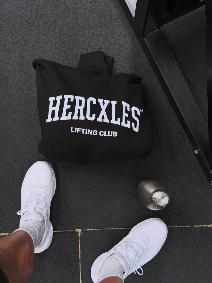 Folded black Lifting Club hoodie with white print beside white trainers and a metal water bottle on a gym floor